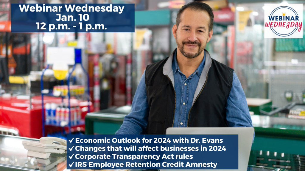 A Hispanic business owner who is male and approximately 45 years old stands at the desk with computer and phone in his hardware store. He is looking forward toward the camera with a smile on his face. Various hardware items are behind him and out of focus.